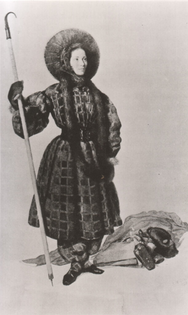 Eine Zeichnung der Bergsteigerin Henriette d'Angeville, die Pluderhosen, einen taillierten Mantel und eine Pelzmütze trägt. In der Hand hält sie einen langen Stab.