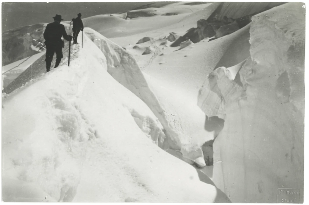 Das Schwarzweißfoto aus dem 19. Jahrhundert zeigt einen tief verschneiten Berg und zwei Bergsteiger in schwarzen Anzügen und Hüten mit Wanderstöcken, die mit einem Seil aneinander befestigt sind.