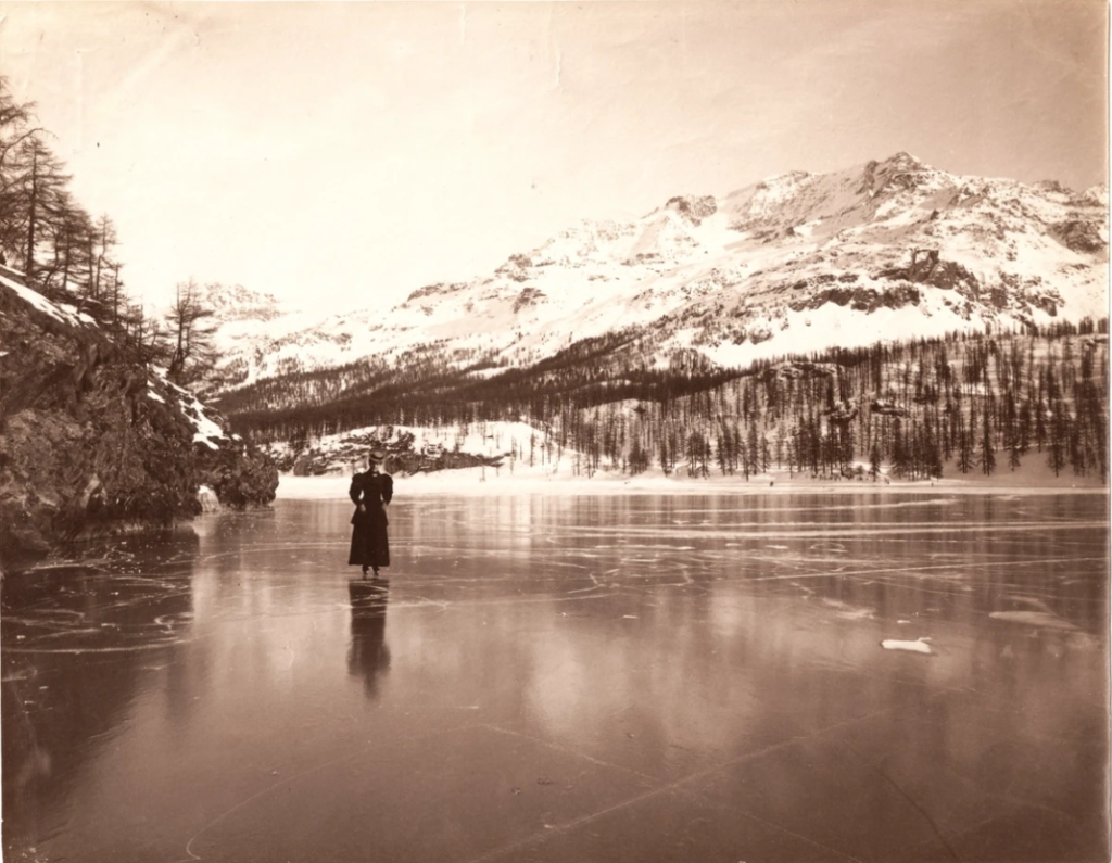 Das sepiafarbene Schwarzweißbild zeigt eine große Eisfläche und im Hintergrund einen schneebedeckten Berg. Auf dem Eis steht eine Frau in schwarzem Kleid und Hut mit Schlittschuhen an den Füßen.