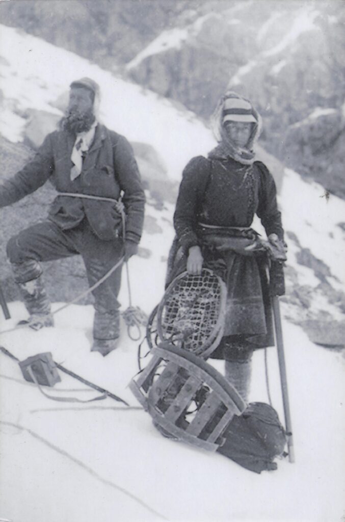 Das Schwarzweißfoto zeigt einen Mann und eine Frau mit Bergsteigerausrüstung. Der Mann blickt in die Ferne. Die Frau hat sich einen Schal um ihren Kopf und Hut geschlungen und hält Schneeschuhe in der Hand.