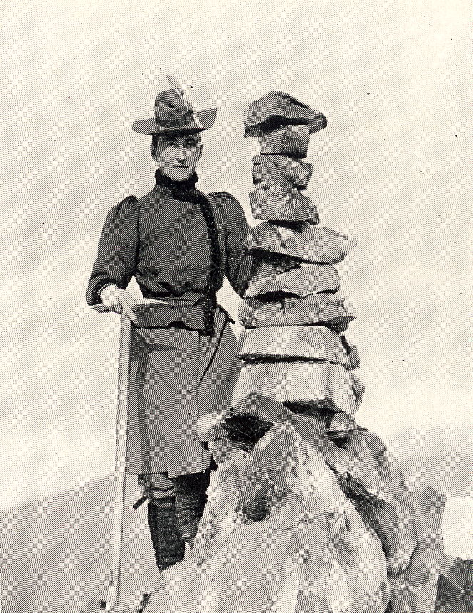 Das Schwarzweißfoto zeigt Elizabeth Main neben einer Steinpyramide auf einem Berggipfel. Sie stützt sich auf einem Stock oder einem Eispickel ab. An ihren Hut hat sie eine Feder geklemmt.