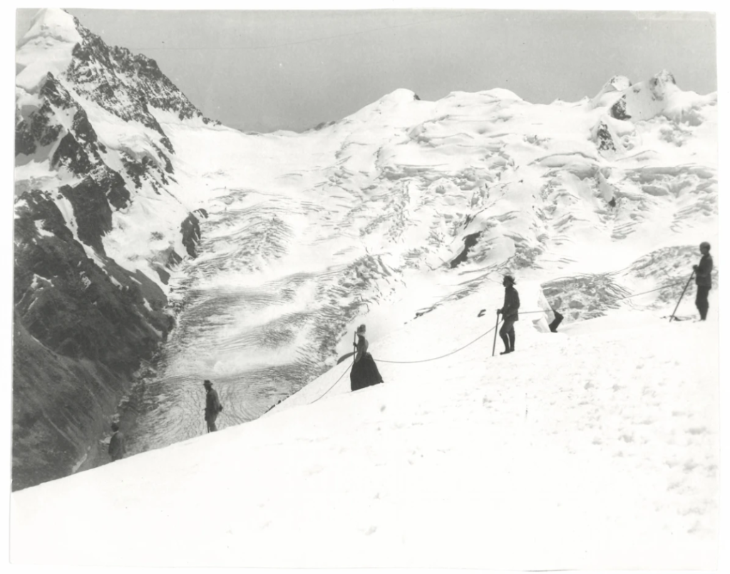 Das Schwarzweißfoto zeigt eine verschneite Berglandschaft. Im Vordergrund sieht man vier Figuren mit Alpenstöcken, die durch ein Seil miteinander verbunden sind.