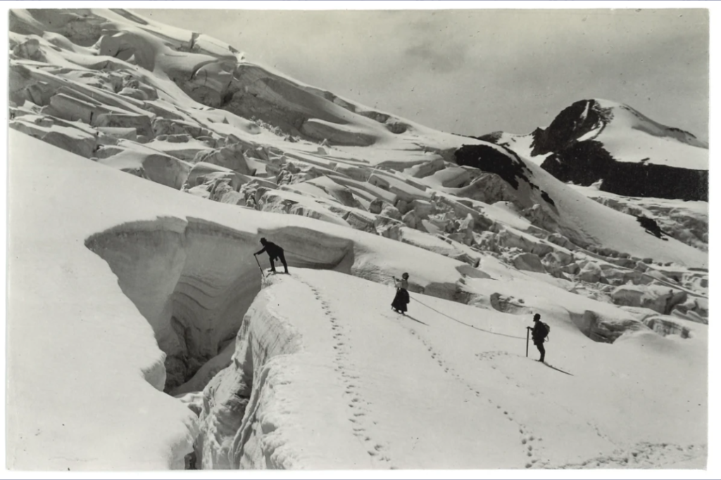 Das Schwarzweißfoto zeigt eine verschneite Berglandschaft. Im Vordergrund klafft eine Gletscherspalte. Ein Mann steht direkt davor, stützt sich auf seinem Stock ab und blickt hinein. Hinter ihm stehen eine Frau und ein Mann, mit denen er über ein Seil zusammengebunden ist.